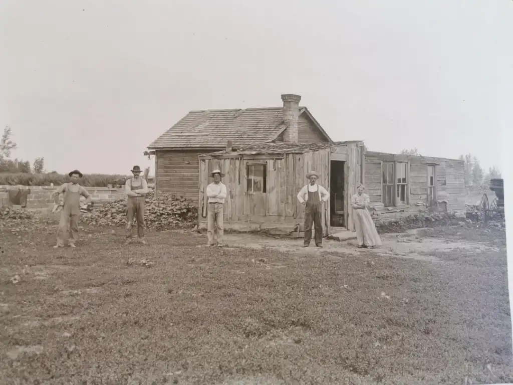 A black and white photo shows five people standing outside a small, rustic wooden building. The structure has a mix of wood and stone walls with a chimney. The group appears to be on a rural homestead with grassy surroundings and some trees.