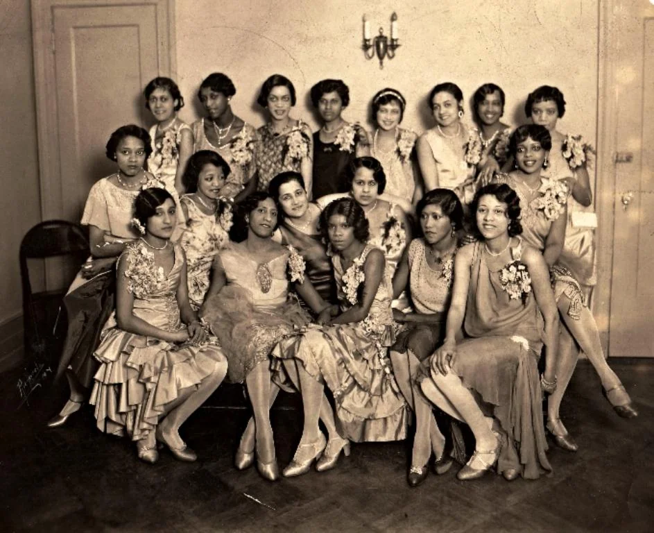 A vintage black and white photo of a group of women dressed in elegant 1920s-style attire. They are posed together in a room, with some seated on a couch and others standing behind. The women wear elaborate dresses with floral details.