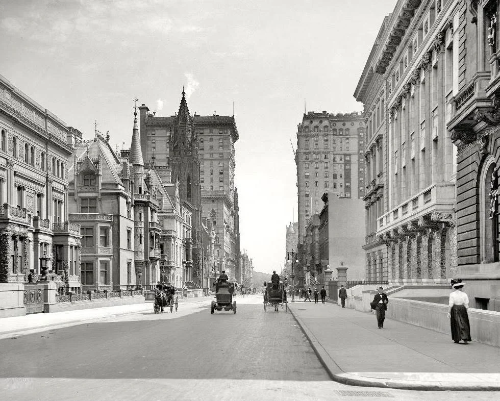 A historic black-and-white photo of a city street lined with ornate buildings from the early 20th century. Horse-drawn carriages and early automobiles travel on the road, while pedestrians in period attire walk on the sidewalk.