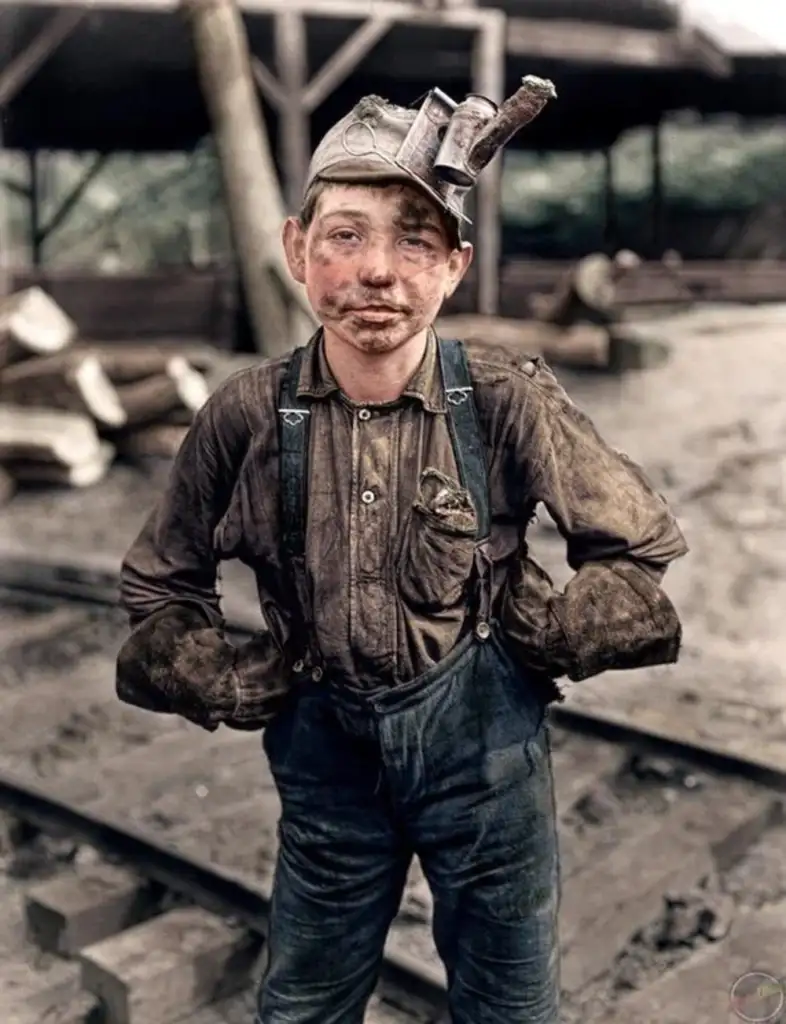 A young coal miner stands on a railway track, wearing a cap with a lamp and gloves. His face and clothes are covered in coal dust. He has his hands on his hips, and the background shows industrial structures and logs.