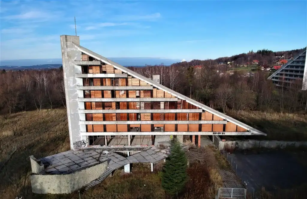 A large, abandoned triangular building with a sloped roof stands in a grassy area. The structure has multiple boarded-up windows and graffiti on its concrete walls. Hills and scattered houses are visible in the background under a clear blue sky.