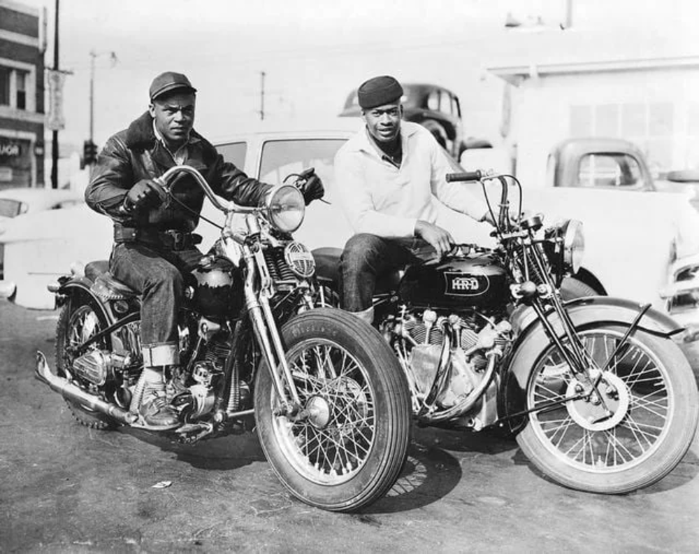 Two men in vintage clothing sit on classic motorcycles parked outdoors. The man on the left wears a leather jacket and boots, while the man on the right is in a cap and light-colored jacket. Cars are visible in the background.