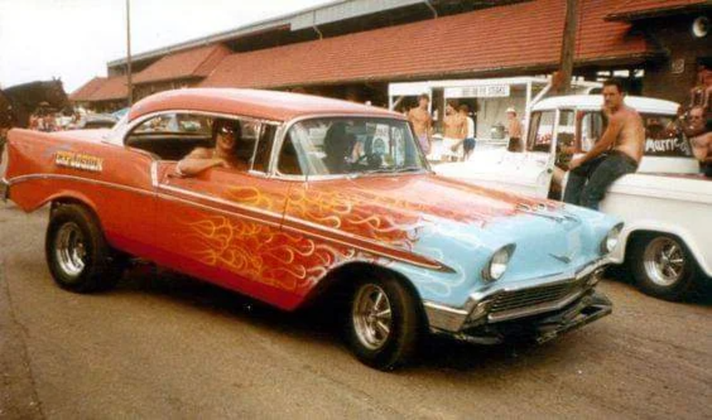 A vintage car with a red and orange flaming paint job is parked on the street. Shirtless men are sitting on the hood of a white truck in the background near a building with a sloping roof.