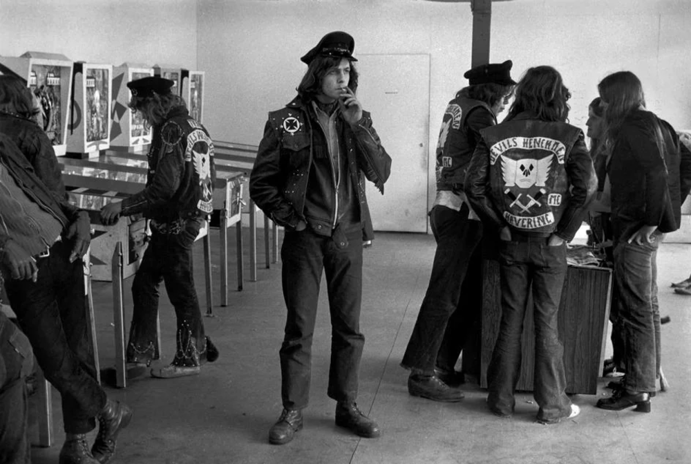 A group of people wearing leather jackets with patches gather indoors, some playing pinball and others in conversation. One person stands in the foreground, smoking, while others have their backs turned. The room has a minimalist design.