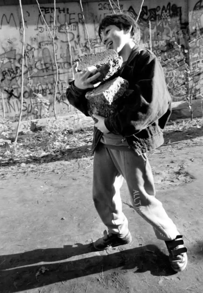 A person wearing a jacket and sweatpants carries three large stones. The background features a graffiti-covered wall and bare trees. The scene is in black and white, and the person appears to be smiling.