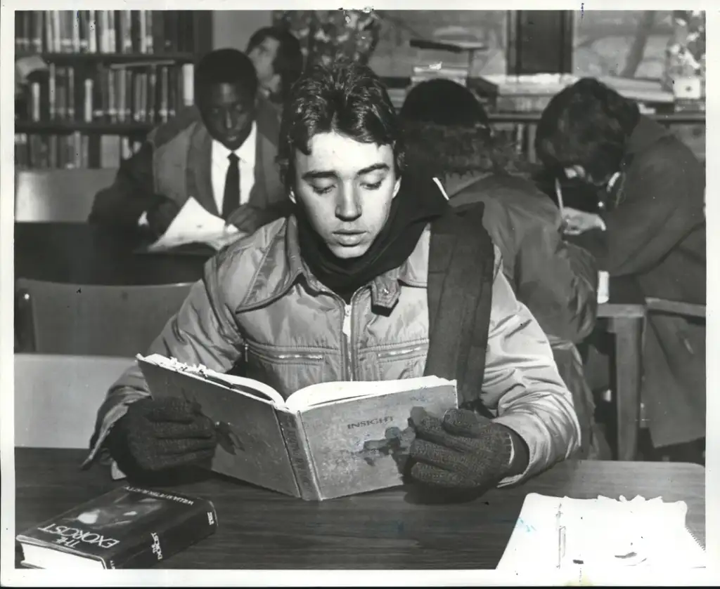 A young person wearing a jacket, scarf, and gloves is focused on reading a book titled "INSIGHT" at a library table. In the background, other people are also engaged in reading and writing activities. Books are scattered on the table.