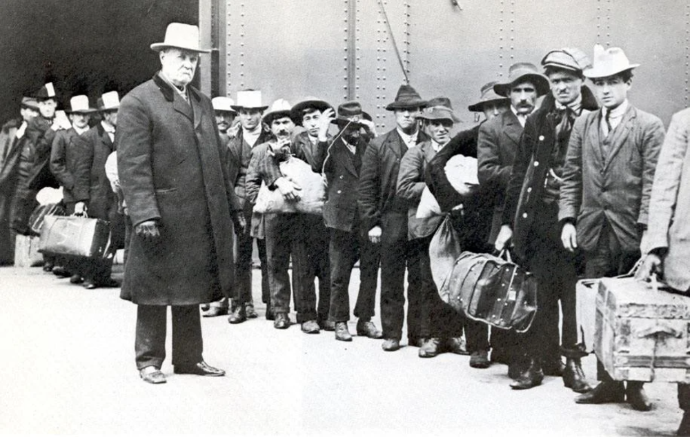 A group of men, dressed in early 20th-century attire, stand in line with luggage, likely immigrants arriving at a port. The men wear hats and coats, and one man stands slightly apart, observing them. A metal wall forms the background.