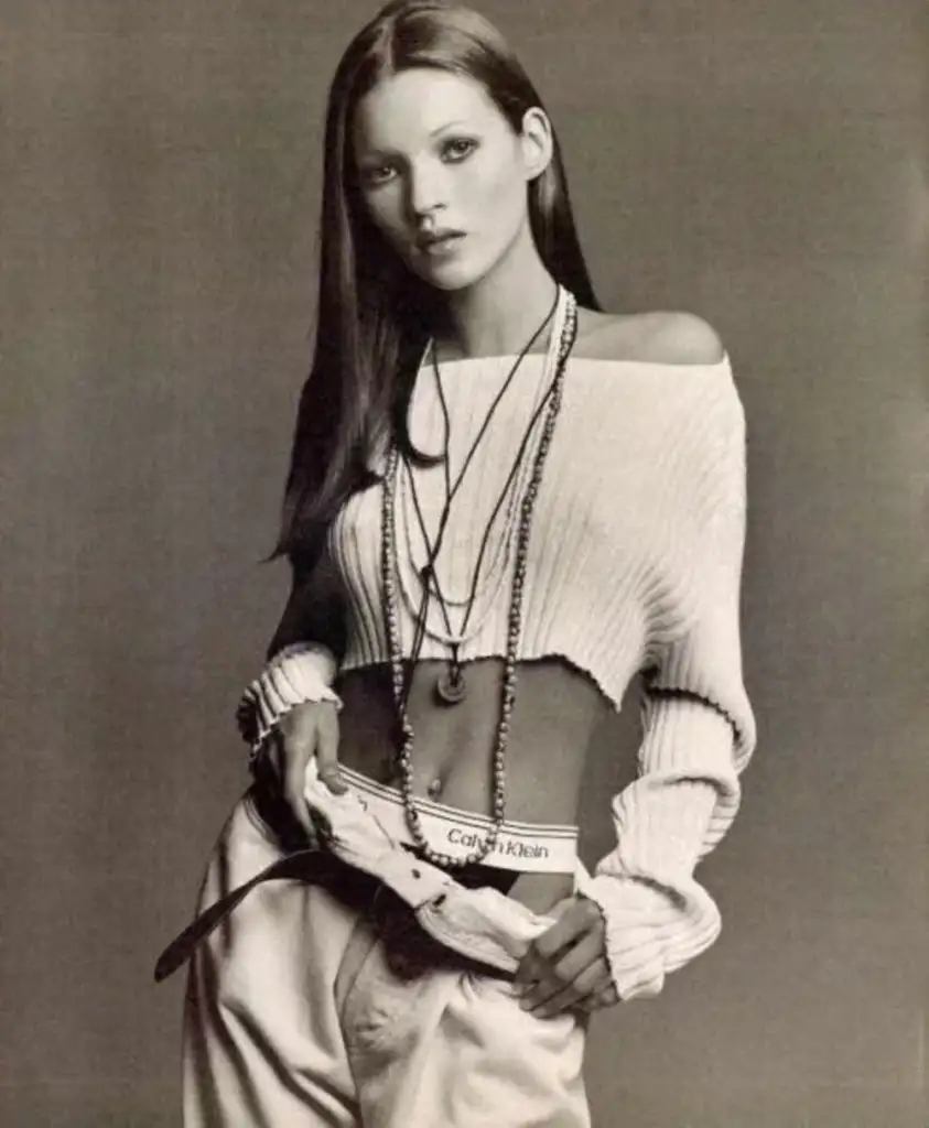 A person with long hair poses in a black-and-white photo. They wear a cropped, off-shoulder sweater and loose pants, revealing Calvin Klein underwear. Several layered necklaces hang around their neck. The image has a classic, vintage feel.