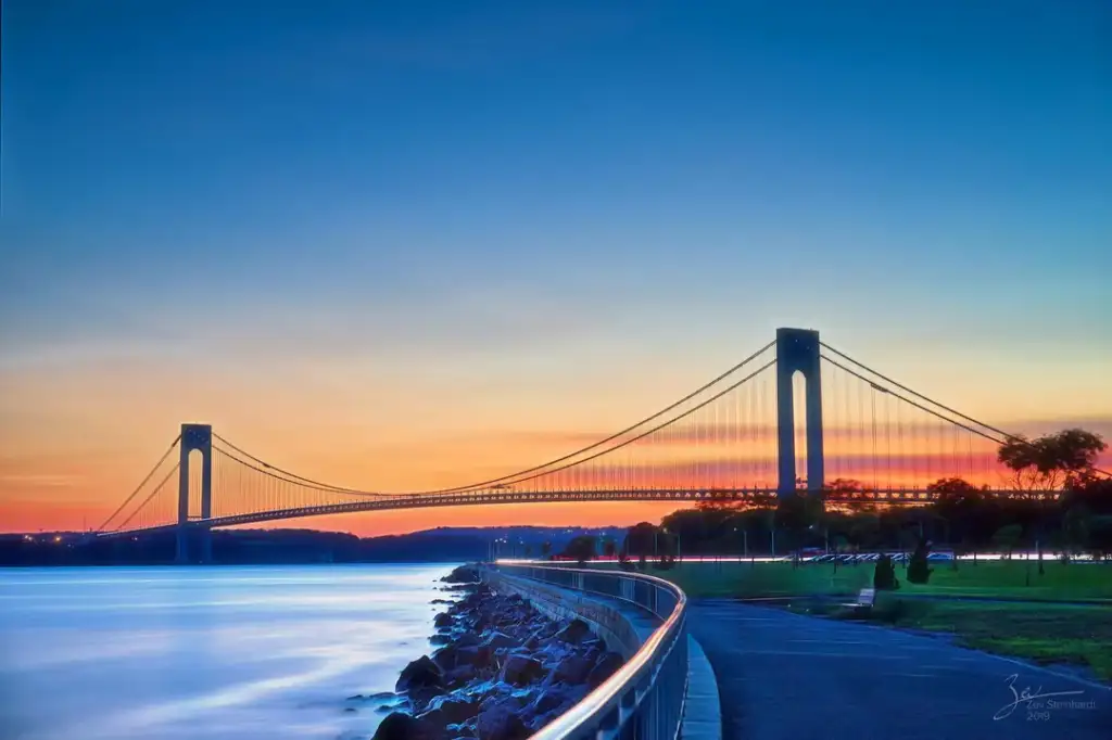 A suspension bridge stretches across a calm body of water at sunset. The sky is painted with vibrant hues of orange, pink, and blue. A paved walkway follows the water's edge, lined with greenery and a few parked cars.