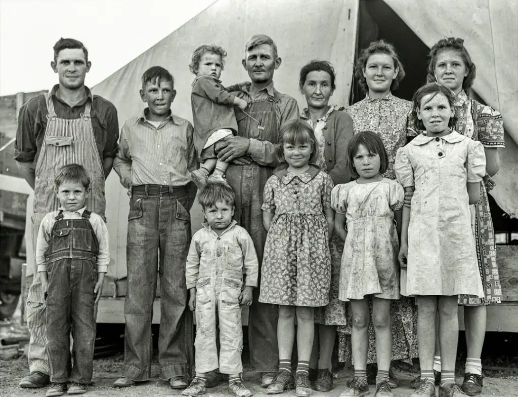 A black and white photo of a family of twelve standing in front of a tent. The group includes adults and children, all dressed in simple, worn clothing. The father holds a toddler. The family appears content, posing closely together.