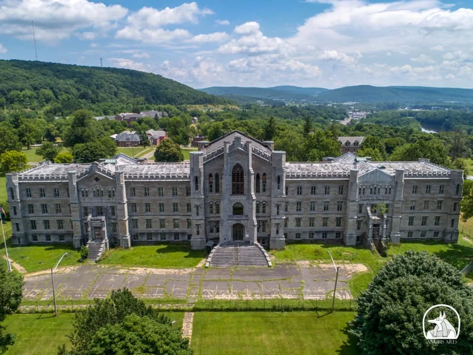 Aerial view of a large, abandoned stone building with Gothic architectural features, surrounded by lush greenery and rolling hills in the background. The sky is partly cloudy, casting soft shadows on the landscape.