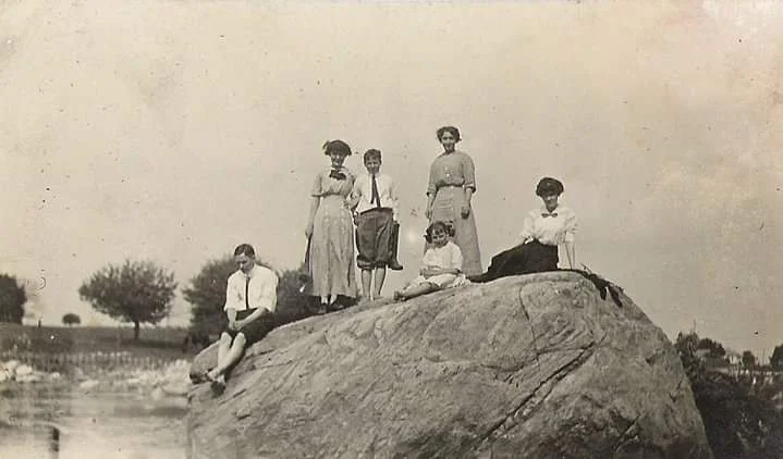 A vintage photo shows six people, three women and three men, standing and sitting on a large rock in an open landscape. They wear early 20th-century attire. The background features trees and an open field under a cloudy sky.