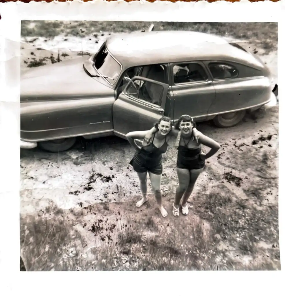 Two women in swimsuits stand on a dirt road with greenery around, smiling at the camera. A vintage car is parked behind them, with both doors open. The photo is black and white and seems to have been taken from above.