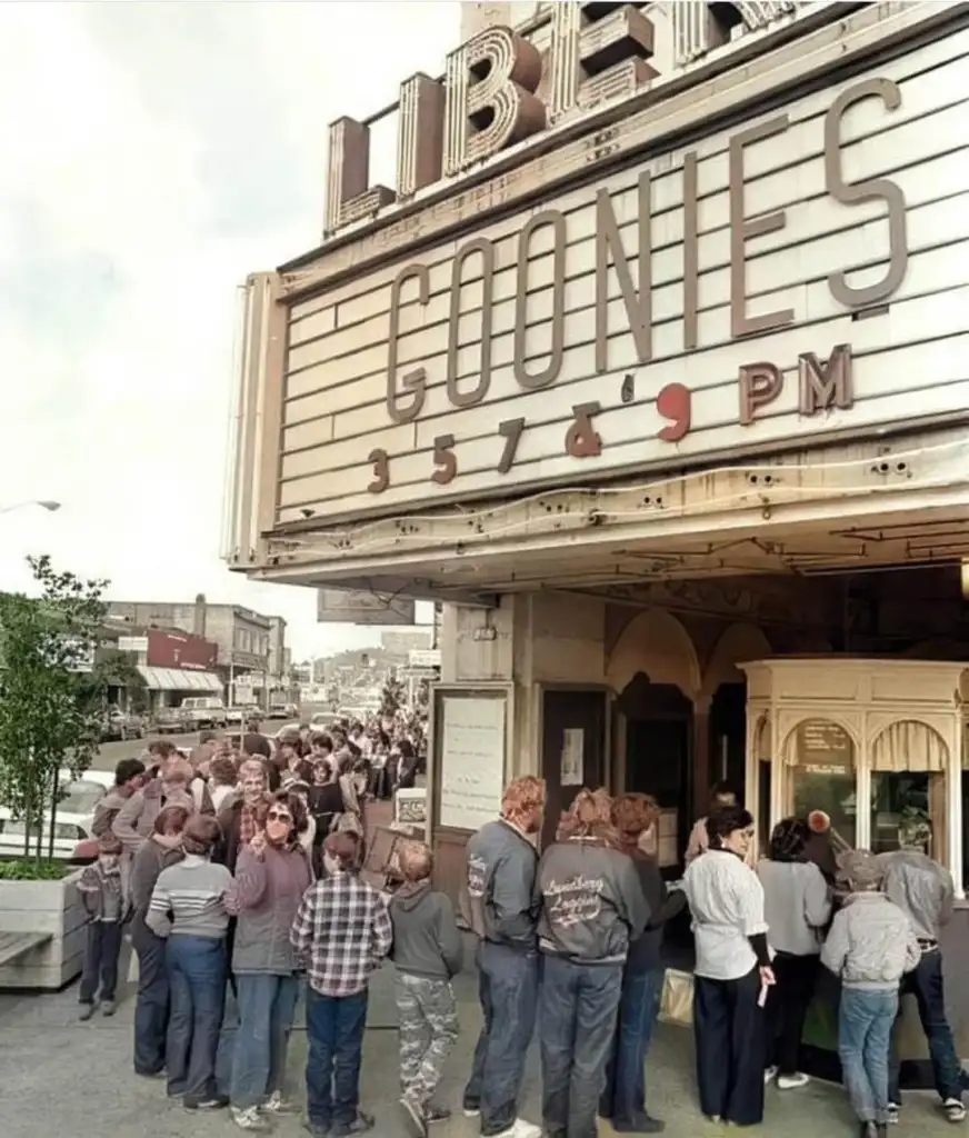 A large crowd of people stand in line outside a theater with a marquee displaying "GOONIES 3, 5, 7 & 9 PM." The theater entrance has a classic design with a ticket booth, and the surrounding street is visible.