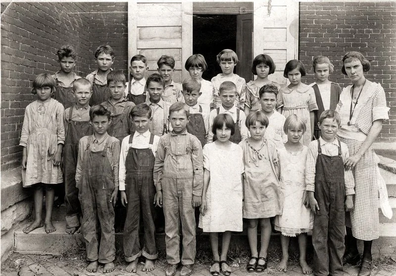 A vintage black and white photo shows a group of children and a woman standing in front of a brick building. The children are wearing overalls and dresses, some are barefoot. They stand in rows with serious expressions. The woman stands to the right.