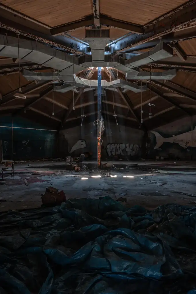 An abandoned circular room with a wooden ceiling, featuring a metal sculpture in the center under a skylight. Sunlight streams in, illuminating scattered debris and a worn tarp. Graffiti-covered walls and a few old chairs add to the derelict atmosphere.