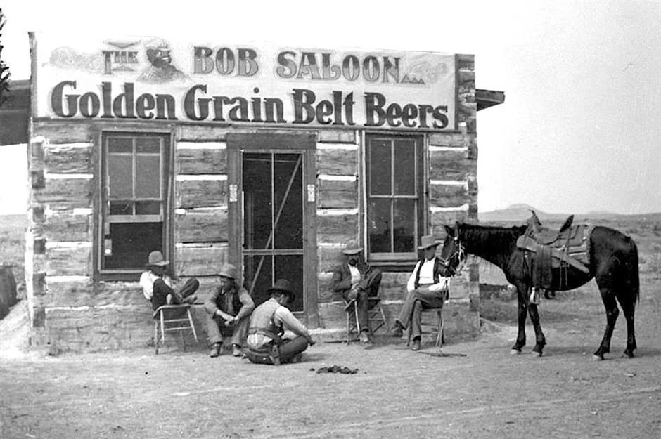 Black-and-white photo of a rustic saloon with a sign reading "Golden Grain Belt Beers." Five people in cowboy hats sit or squat outside, with a horse tied nearby. The building is made of logs, set in a barren landscape.