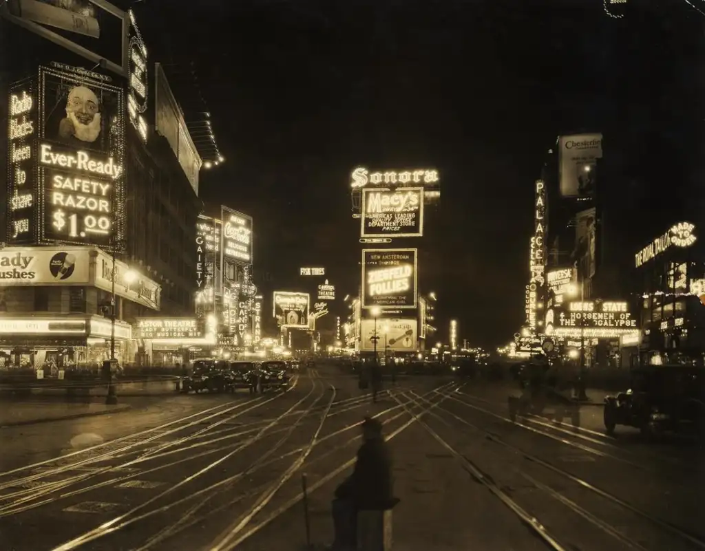 A vintage nighttime scene of Times Square, New York City. Bright neon signs and billboards illuminate the streets, advertising products like Ever-Ready razors and Macy's. Tram tracks and a few cars are visible, with a person crossing the street.