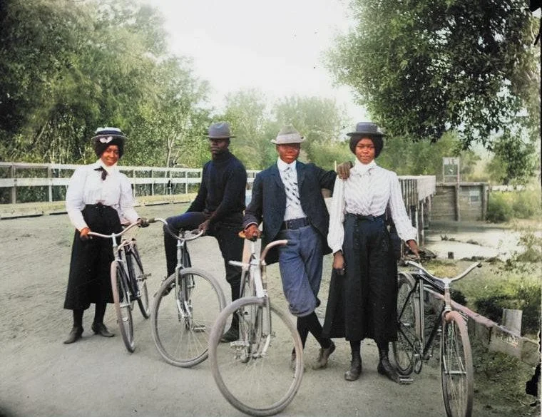 Four people stand on a bridge with bicycles, wearing early 1900s attire, including hats and long skirts. They appear relaxed and are surrounded by trees.