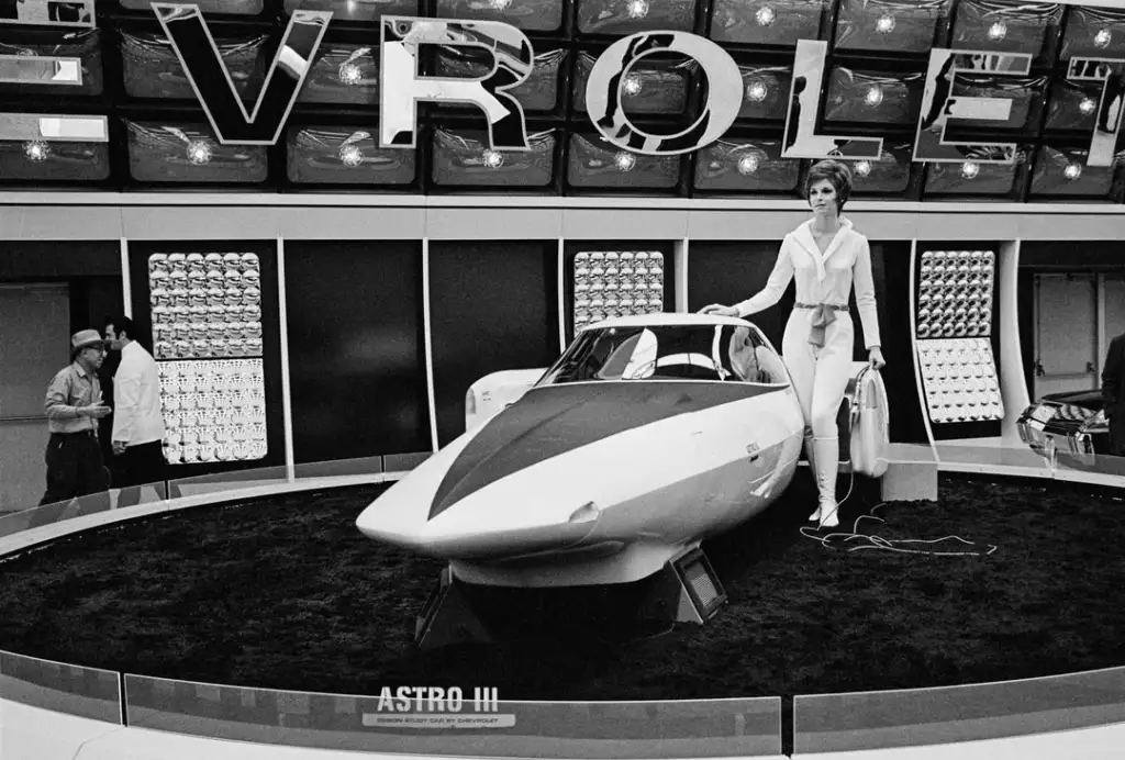 A woman in a futuristic outfit stands next to the sleek, aerodynamic Chevrolet Astro III concept car at an auto show. The background features displays with illuminated panels and the Chevrolet logo.