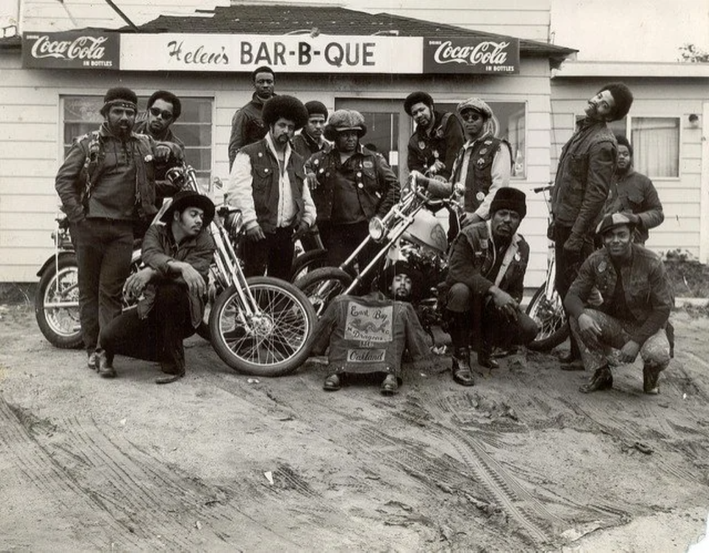 A group of people posing with motorcycles in front of a building labeled "Helen's BAR-B-QUE" with Coca-Cola signage. They are dressed in leather jackets and caps, and some are sitting on the bikes while others stand around. The setting looks vintage.