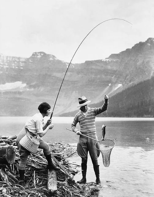 Two people fishing by a lake against a mountainous backdrop. One, holding a fishing rod, has just caught a fish. The other person stands nearby with a fishing net. Both are dressed in early 20th-century attire. Logs and water surround them.