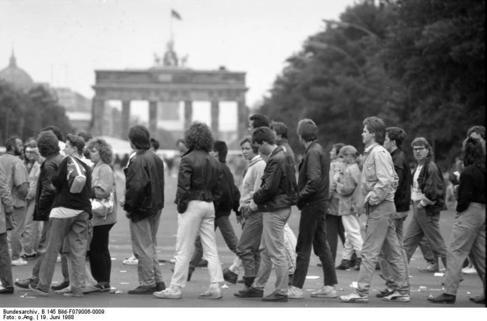 A black-and-white photo of a group of people walking and gathering in front of the Brandenburg Gate in Berlin. The scene appears casual, with individuals dressed in 1980s style clothing.