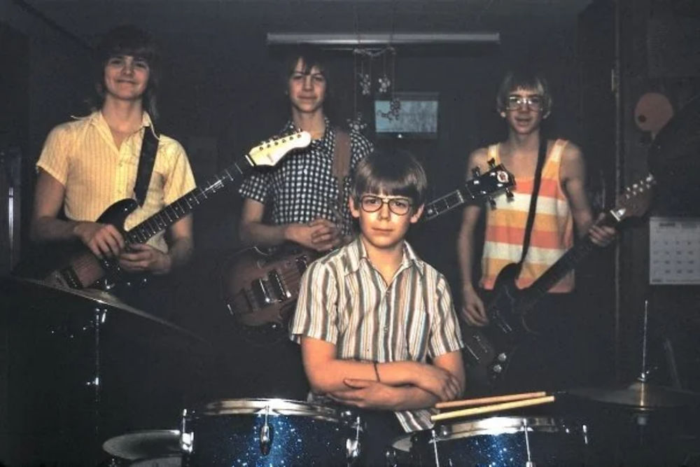 A young boy sits at a drum set with drumsticks in hand, while three other boys stand behind him holding guitars. They are in a dimly lit room, smiling at the camera. One boy is in a sleeveless striped shirt, and another wears glasses.