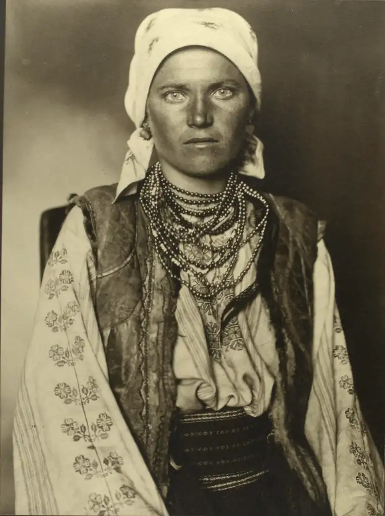 A sepia-toned portrait of a woman wearing traditional Eastern European attire. She is adorned with layered necklaces, an embroidered blouse, a vest, and a headscarf. Her expression is serious, and she looks directly at the camera.