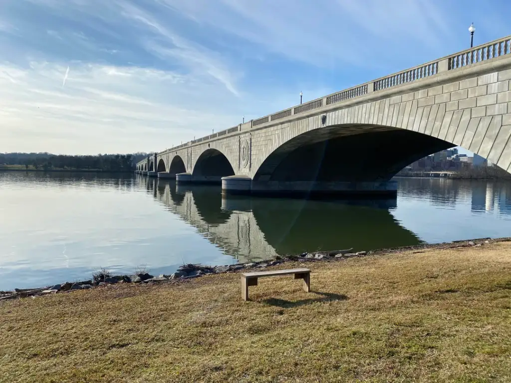 A stone bridge with multiple arches spans across a wide, calm river. The sky is clear with a few wispy clouds. A lone wooden bench is placed on the grassy riverbank in the foreground, casting a shadow on the ground.