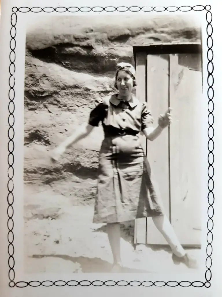 Black and white photo of a woman in a dress, standing in front of a wooden door set in a rocky wall. She has one arm raised and is smiling. The image is framed with a decorative border.