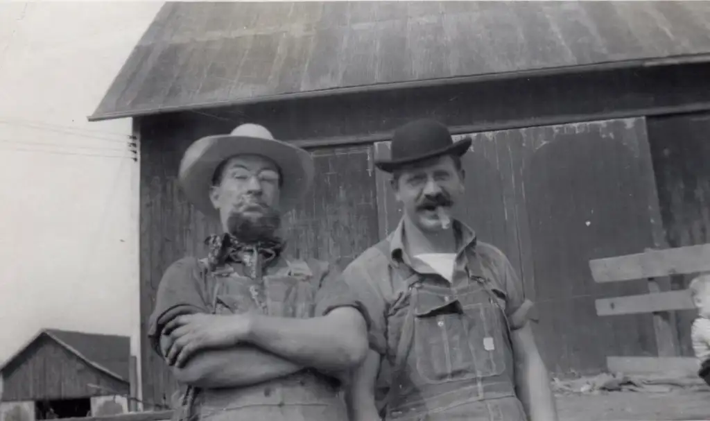 Two men stand in front of a wooden barn. Both wear overalls; one has a large brim hat and beard, the other a bowler hat and mustache. A small child is partially visible on the right. The image has a vintage, black-and-white appearance.