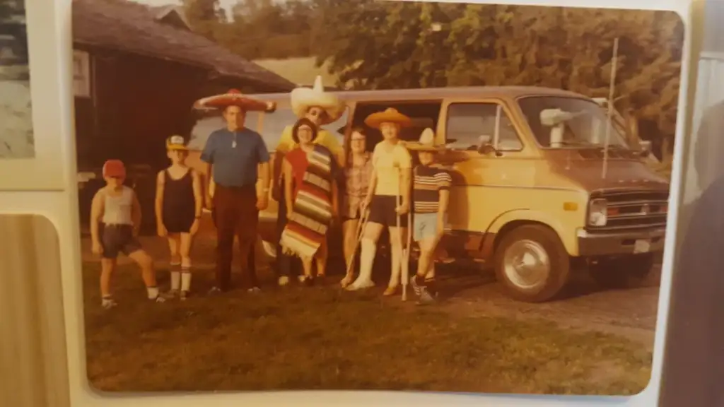 A group of seven people stands in front of a brown van, outdoors. They are wearing a mix of sombreros, ponchos, and casual summer clothing. Trees and a house are in the background, suggesting a rural setting.