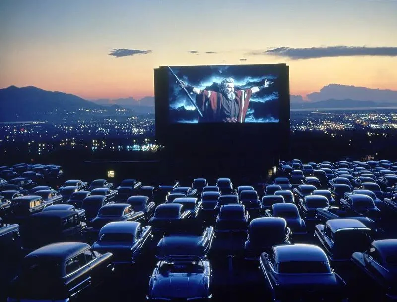 A drive-in theater at dusk with rows of vintage cars facing a large screen showing a scene from a film featuring a person holding a staff with arms raised. City lights and mountains are visible in the background under a colorful sky.