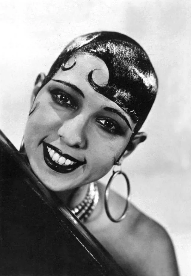 Black and white photo of a woman with a close-cropped hairstyle and styled waves. She smiles broadly, wearing large hoop earrings and a multi-tiered necklace. Her head is tilted slightly to the right.