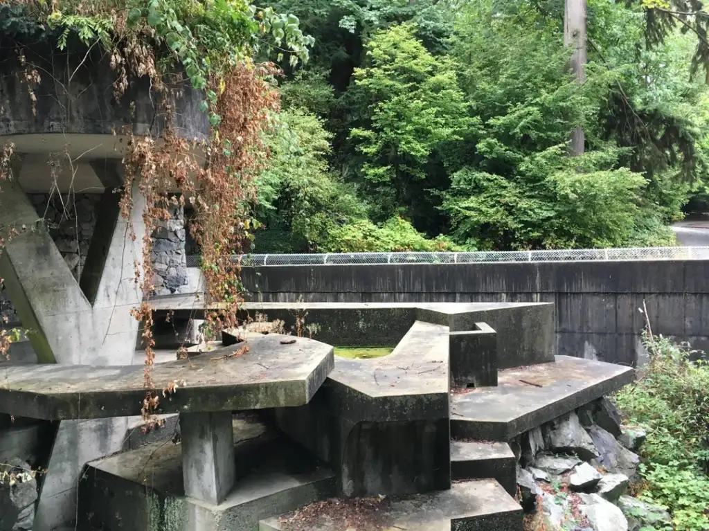 Concrete platforms and structures with angular designs are surrounded by overgrown bushes and trees. The scene includes a wall and a street in the background, flanked by dense greenery.