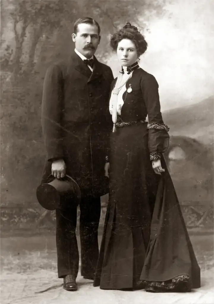 A vintage black and white portrait of a man and a woman from the late 19th or early 20th century. The man in a dark suit holds a bowler hat. The woman wears a long, elegant dress with lace details, standing beside him.