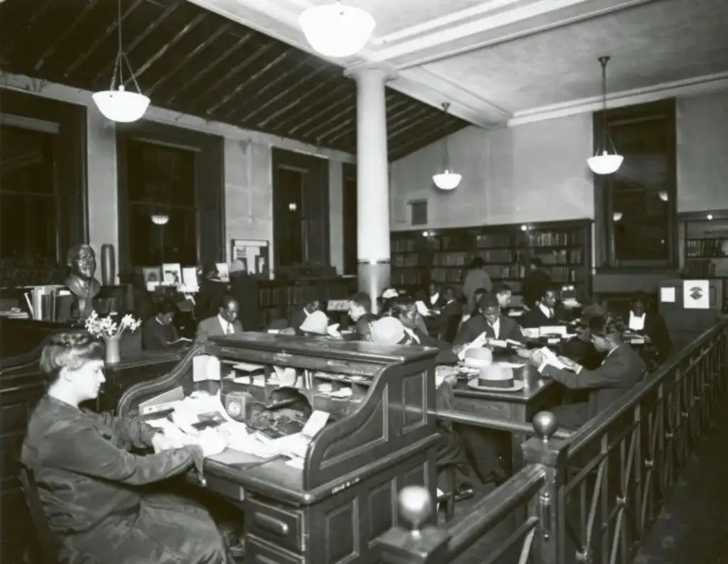 A black-and-white photo of a busy library with people sitting at desks reading and studying. Shelves of books line the walls, and a bust is displayed in the corner. Overhead, several round lights illuminate the spacious room.