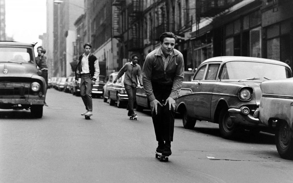 Black and white photo of four people skateboarding down a city street lined with vintage cars. The leader leans forward, and the others follow in casual attire. Urban buildings and a truck are visible. The scene captures a sense of adventure and freedom.