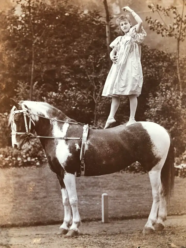 A young girl in a dress stands balanced on a large, piebald horse in an outdoor setting. She is holding one arm up gracefully and gazing upward. Trees and foliage are visible in the background. The image has a vintage sepia tone.