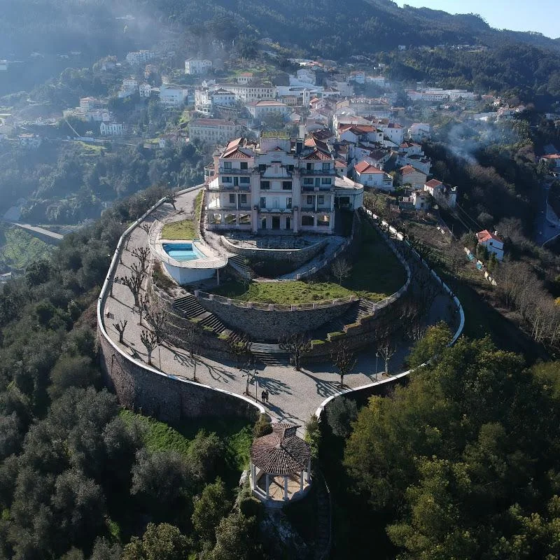 Aerial view of a large, multi-story building on a hill surrounded by tiered stone walls and a circular road. There's a swimming pool nearby, and the background shows a town nestled in a forested mountainous area. Smoke rises from parts of the town.