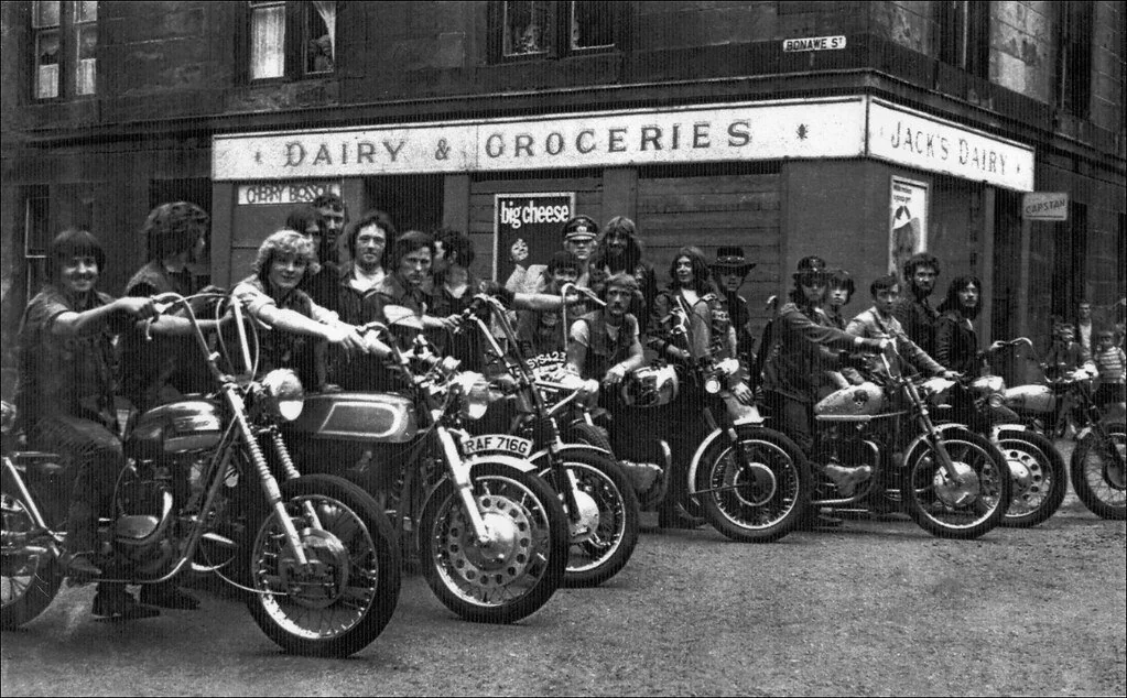 A group of people sitting on motorcycles in front of a store with a "Dairy & Groceries" sign. The scene appears vintage, with the riders dressed in casual, retro attire, and the motorcycles parked in a line on the street.