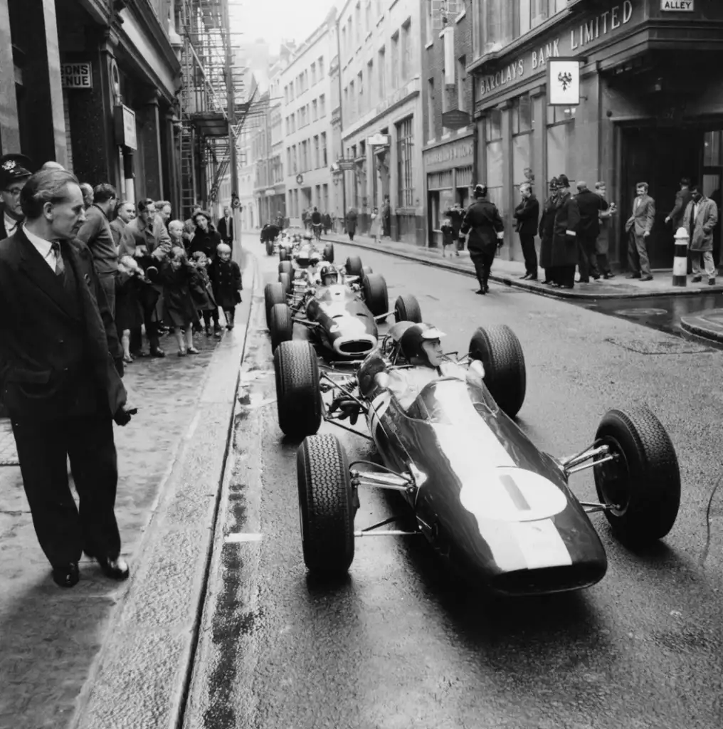 A row of vintage race cars is parked on a narrow city street. People, including children, stand on the sidewalk watching. Buildings and a bank are visible in the background. A man in a suit stands in the foreground, facing the cars.