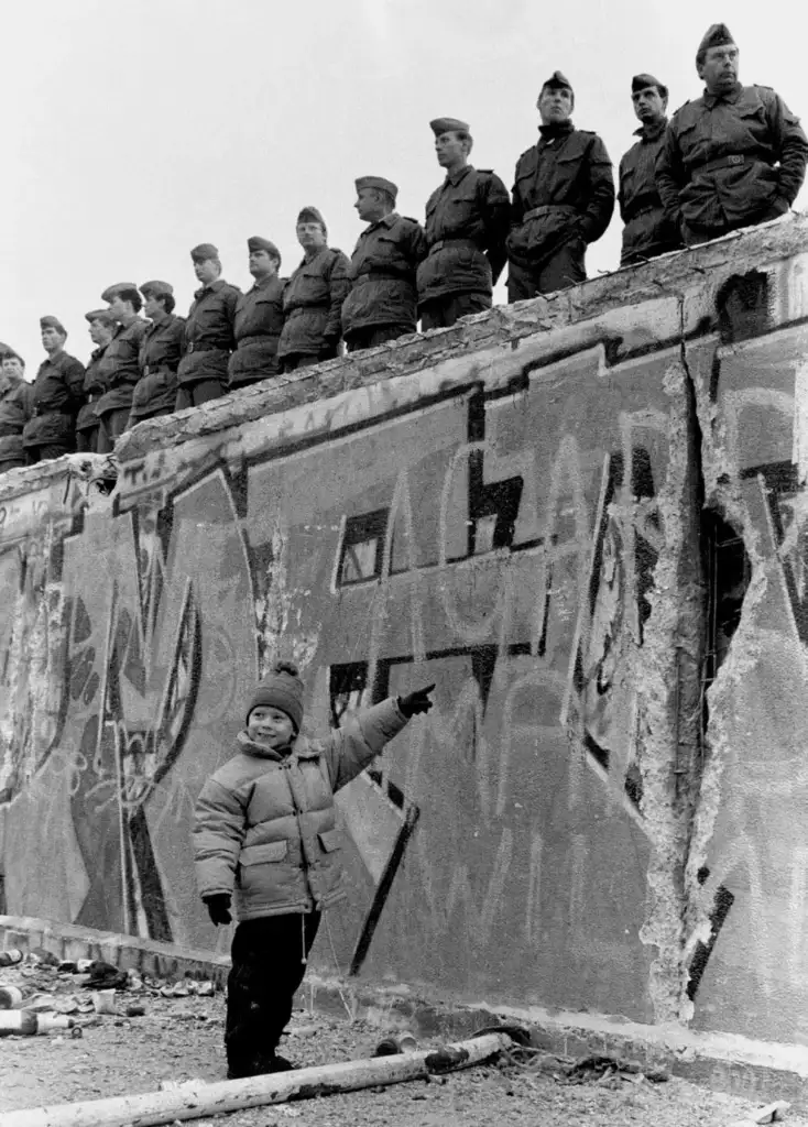 A child in winter clothing points at a graffiti-covered wall with soldiers standing on top. The wall shows signs of decay, with debris scattered on the ground. The soldiers are wearing uniforms and berets, standing in a row.