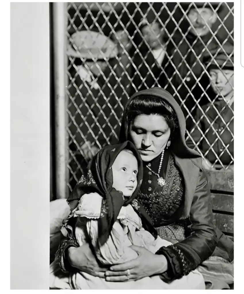 A woman in traditional clothing sits holding a young child on her lap. Both wear headscarves. They are in front of a chain-link fence, with several people visible in the background. The photo is in black and white.