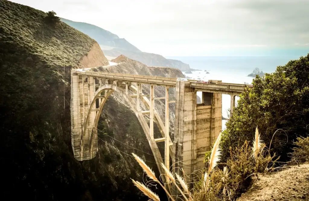 The image shows a tall, arched concrete bridge spanning a deep canyon. It is surrounded by rugged cliffs and overlooks a scenic coastline with the ocean in the distance. The bridge and landscape are bathed in warm, golden light.