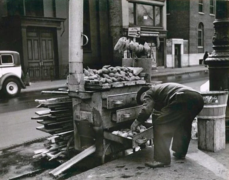 A street vendor leans over a cart filled with sweet potatoes, adjusting a basket. The cart is on a city sidewalk, with buildings, a vintage car, and a lamppost in the background. Bags are piled on top of the cart.