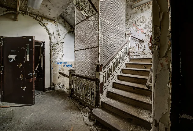 An abandoned, decaying staircase with peeling paint and cracked walls. A door is open on the left, and graffiti marks the rough, worn surfaces. The setting suggests neglect and age, with debris scattered on the floor.