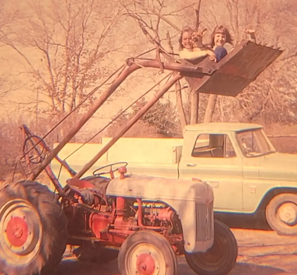 Two people sit in the raised bucket of a red tractor. They are smiling and waving. In the background, there is a vintage green pickup truck and leafless trees. The image appears to have a faded, nostalgic look.