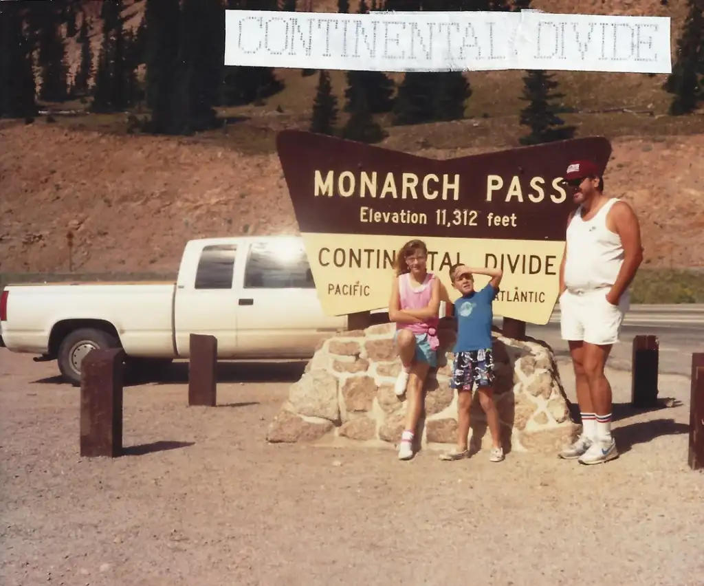 Three people are posing in front of the Monarch Pass sign, which reads "Elevation 11,312 feet" and "Continental Divide." A white pickup truck is parked nearby. The setting is mountainous with trees in the background.